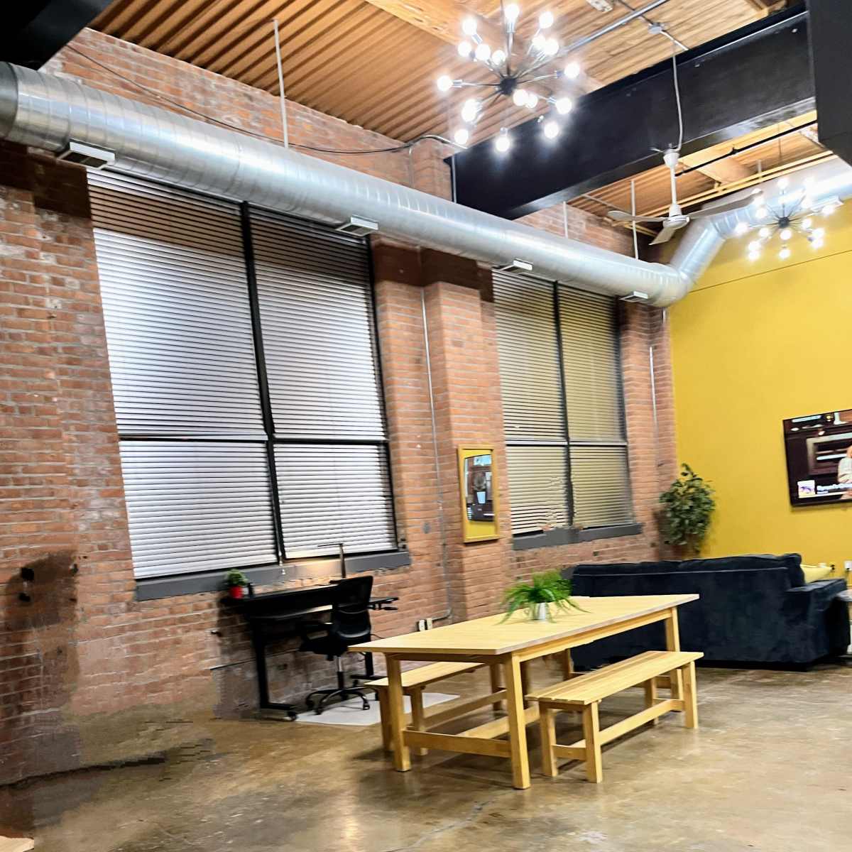 A spacious industrial-style room with exposed brick walls, large windows, and a wooden table surrounded by benches, along with a chair and a green plant on the table.