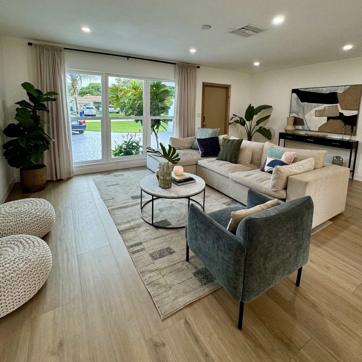 A modern living room features a beige sofa, a circular coffee table, and a large window overlooking a garden area.