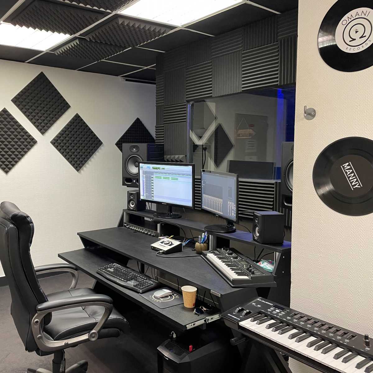 A soundproofed music studio featuring a black desk with computer monitors, audio equipment, and two ergonomic chairs.
