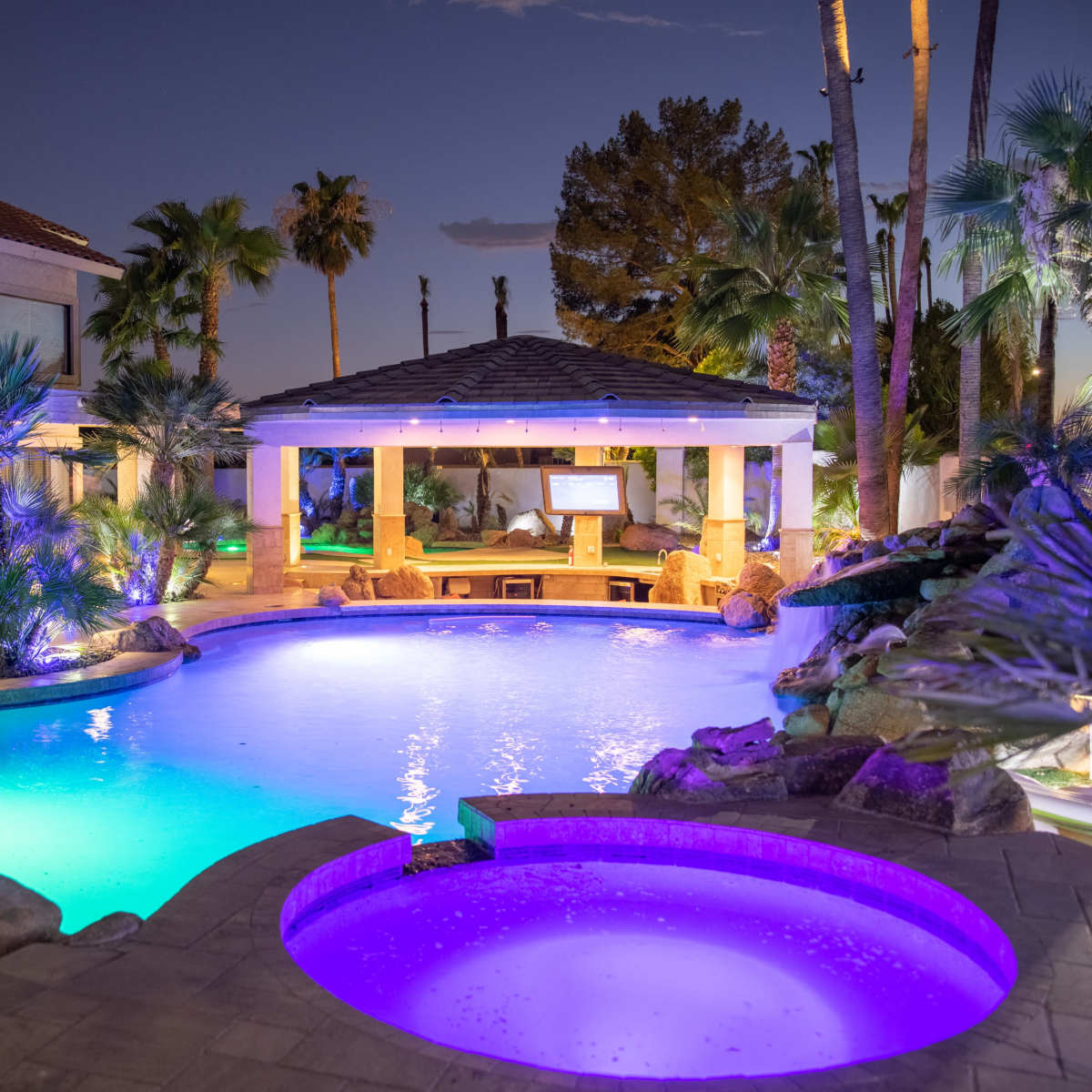 A well-lit backyard pool area at night, featuring a swimming pool, a spa, and tropical landscaping with palm trees.