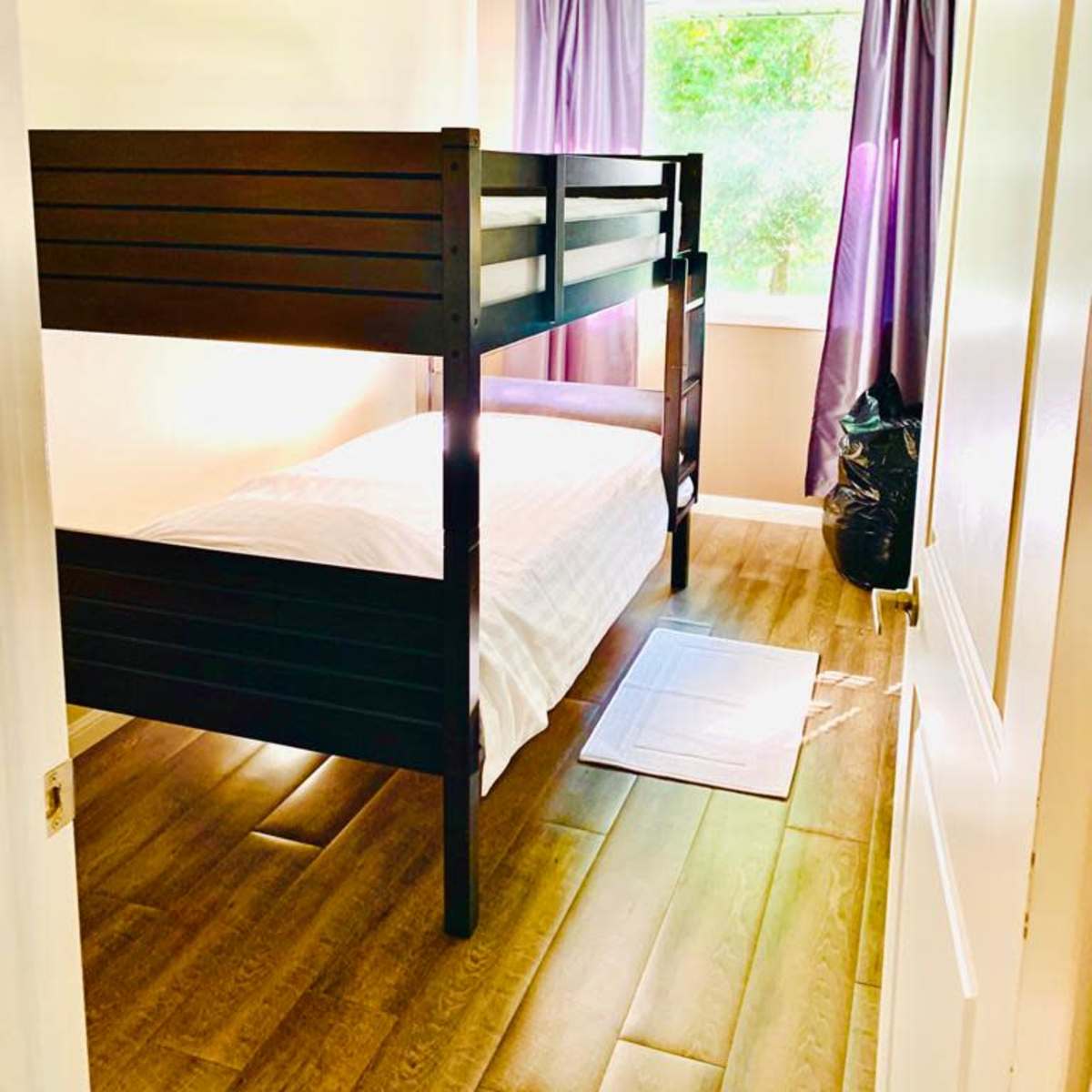 A small bedroom featuring a black bunk bed, a single bed on the bottom, a window with purple curtains, and a wooden floor.
