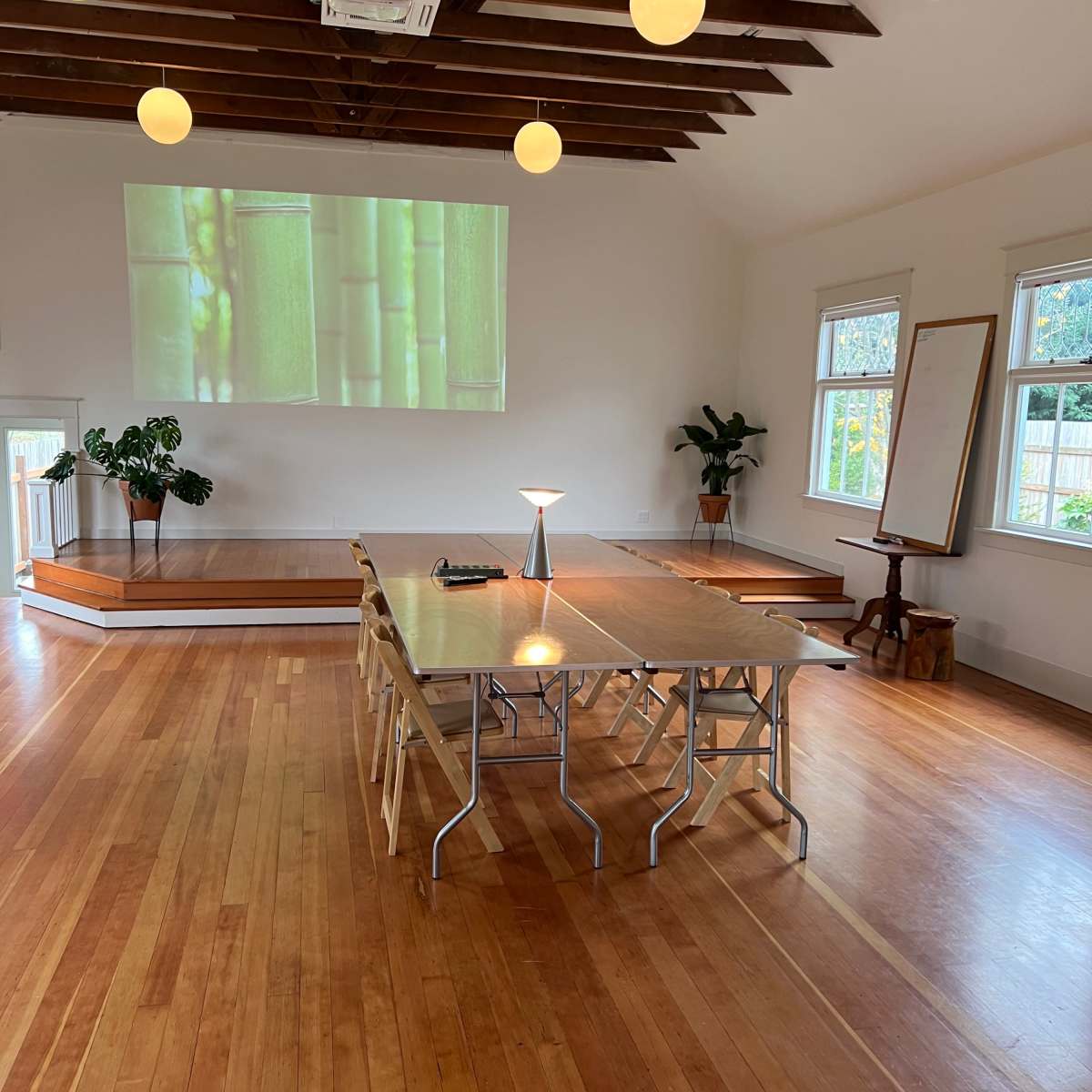 A spacious, well-lit room with wooden floors, featuring a large table set for a meeting, a projector displaying bamboo on the wall, and potted plants in the corners.