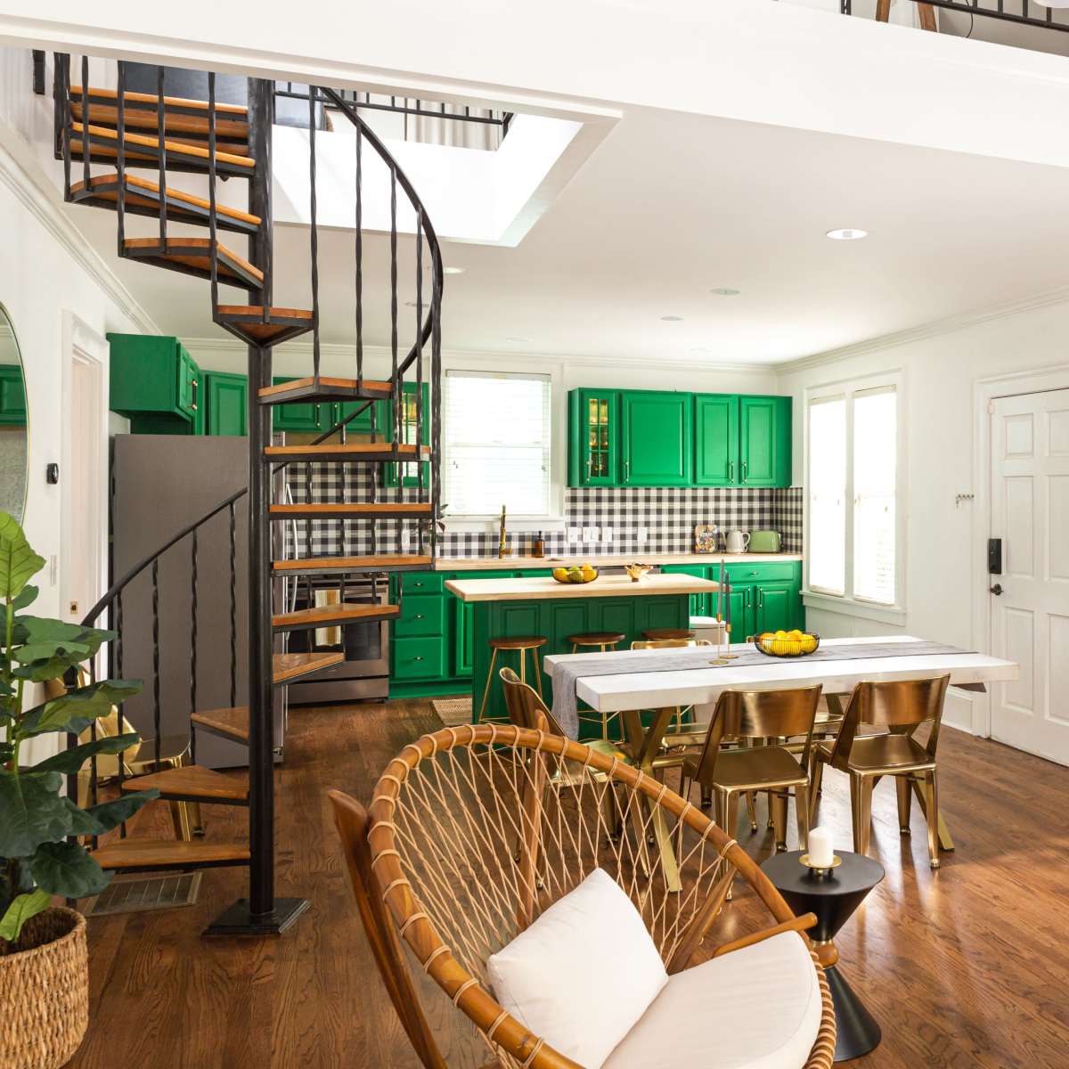 A modern open-concept kitchen and living area featuring green cabinets, a spiral staircase, and a dining table surrounded by various furnishings.