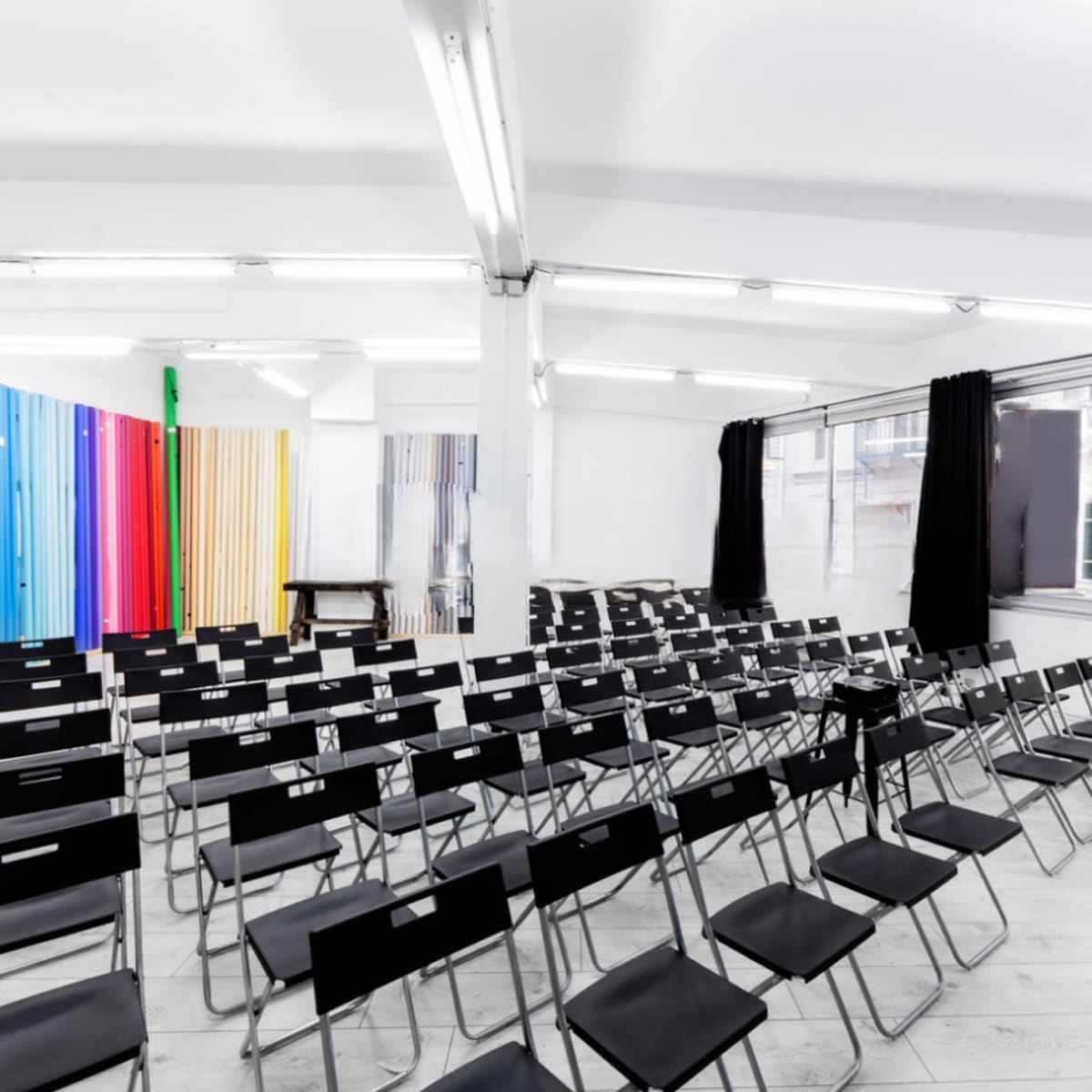 An empty room filled with rows of black folding chairs facing a blank wall, with colorful panels in the background.