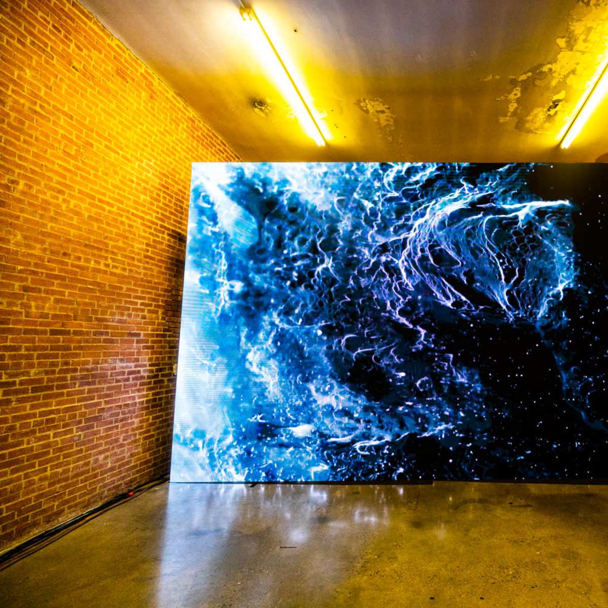 A large digital screen displaying a vibrant abstract visual is installed on a brick wall in a dimly lit room with exposed lighting fixtures.