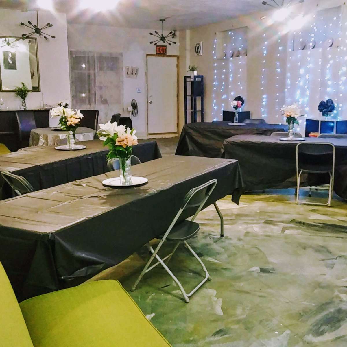 A room set up for an event, featuring several tables covered with black tablecloths and floral centerpieces, with soft lighting and decorative fairy lights in the background.