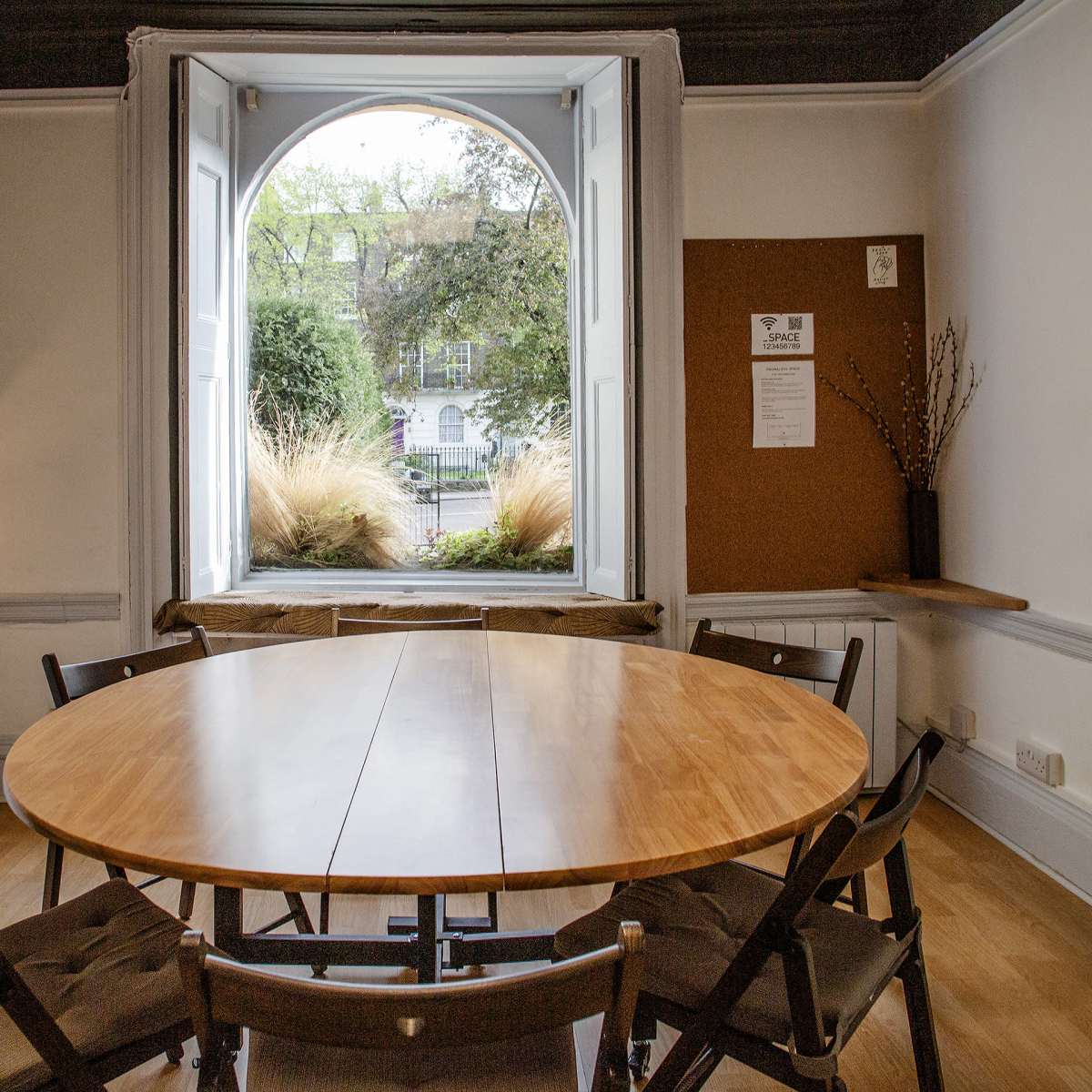 A round wooden table surrounded by chairs is positioned in a well-lit room with a large circular window overlooking a garden.