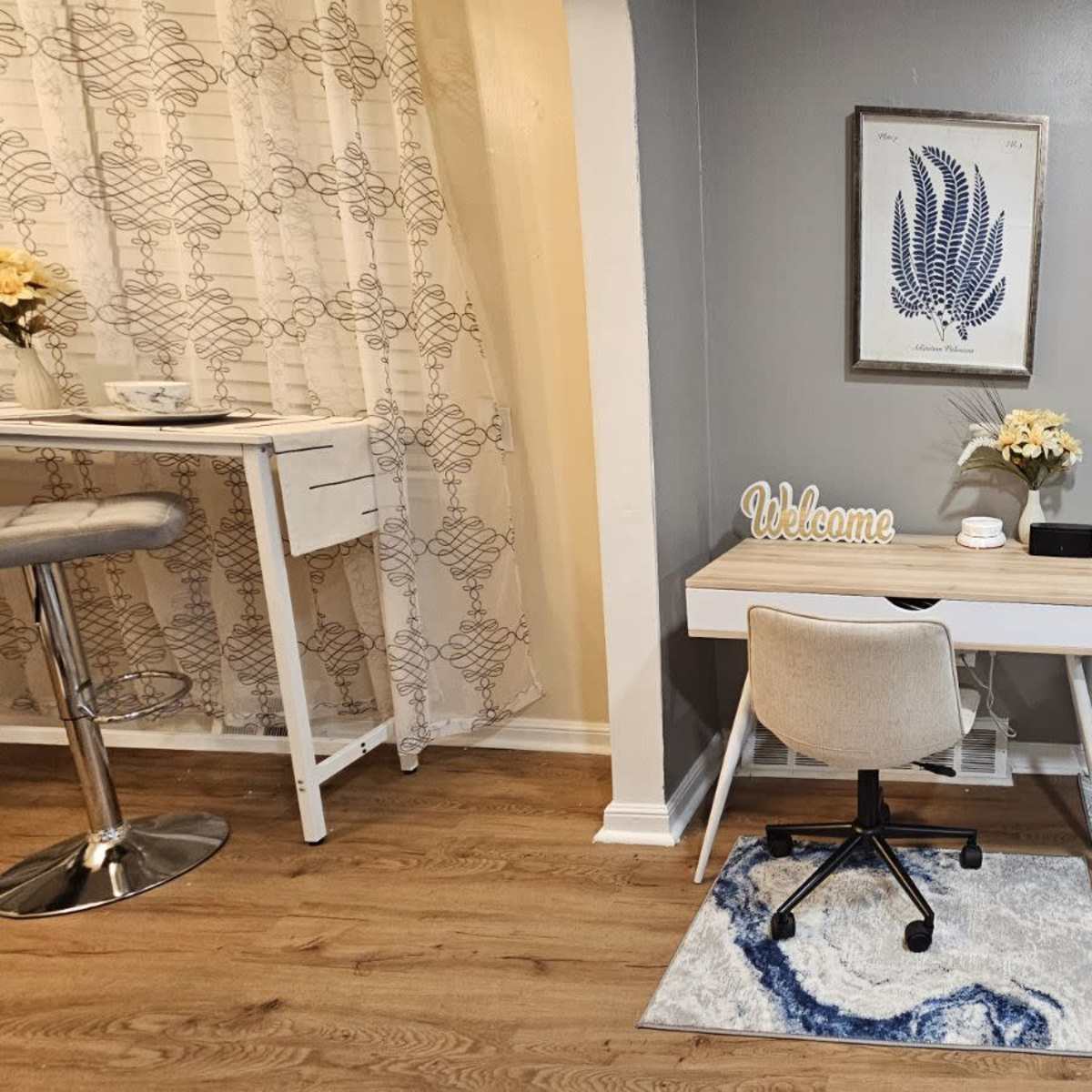 A modern living space featuring a blue sofa, a small desk with a chair, a wall clock, and decorative elements like a framed artwork and flowers.