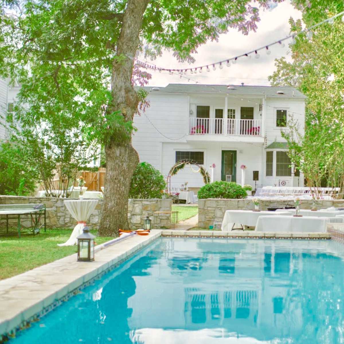 A clear swimming pool reflects a two-story house surrounded by greenery and outdoor seating areas for gatherings.