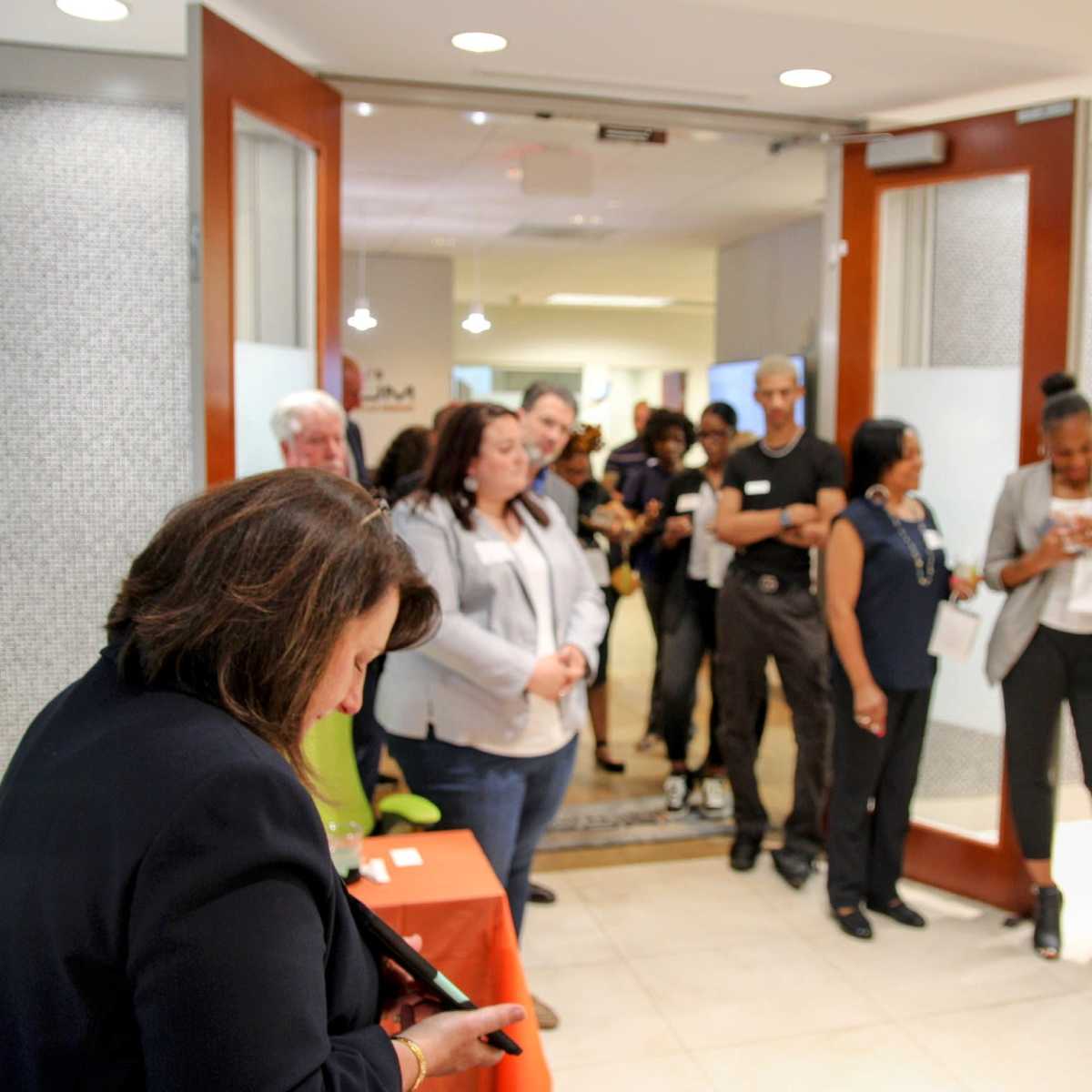 A group of people stands in line inside a building, with some individuals interacting with their smartphones and others engaged in conversation.