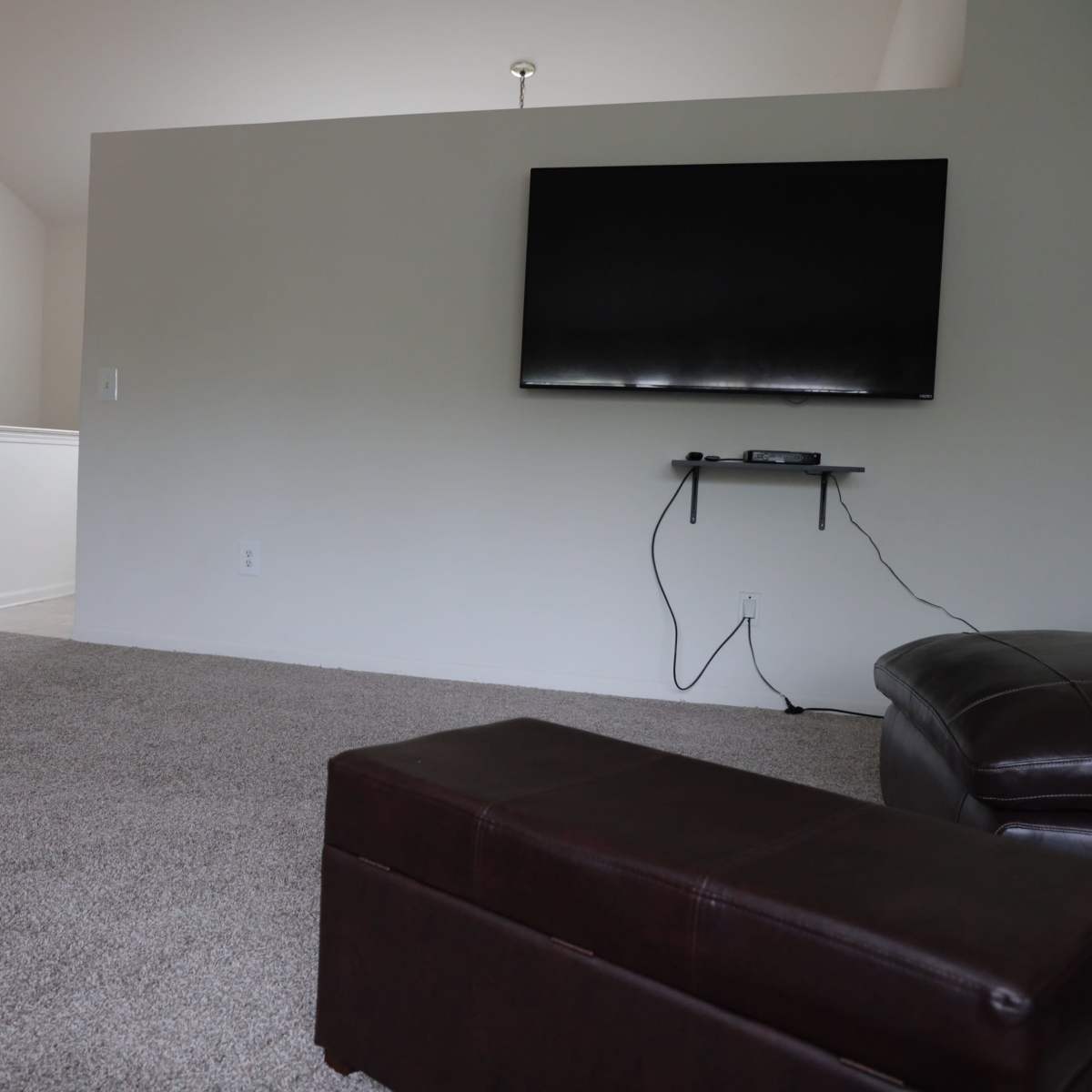 A living room with carpeted flooring, a wall-mounted television, and a brown leather ottoman.