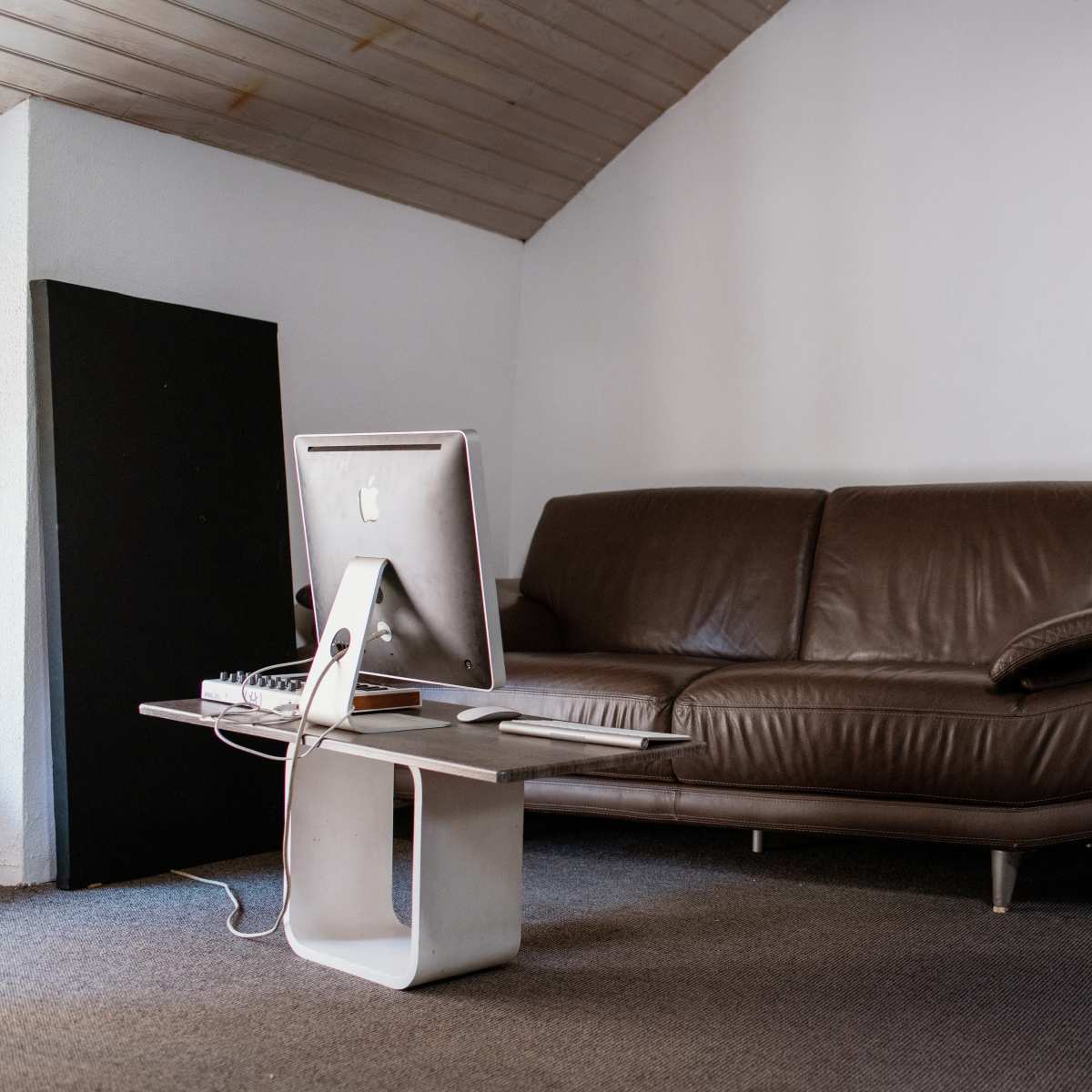 A brown couch is positioned in a room with a computer on a table and a guitar leaning against the wall.