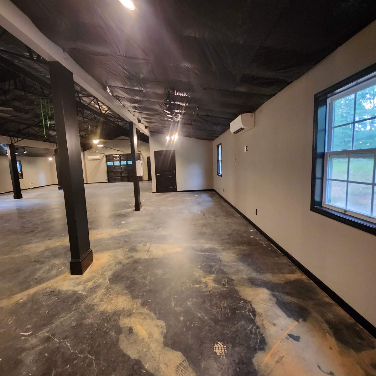 An empty interior space with concrete flooring, black ceiling tiles, and large windows providing natural light.