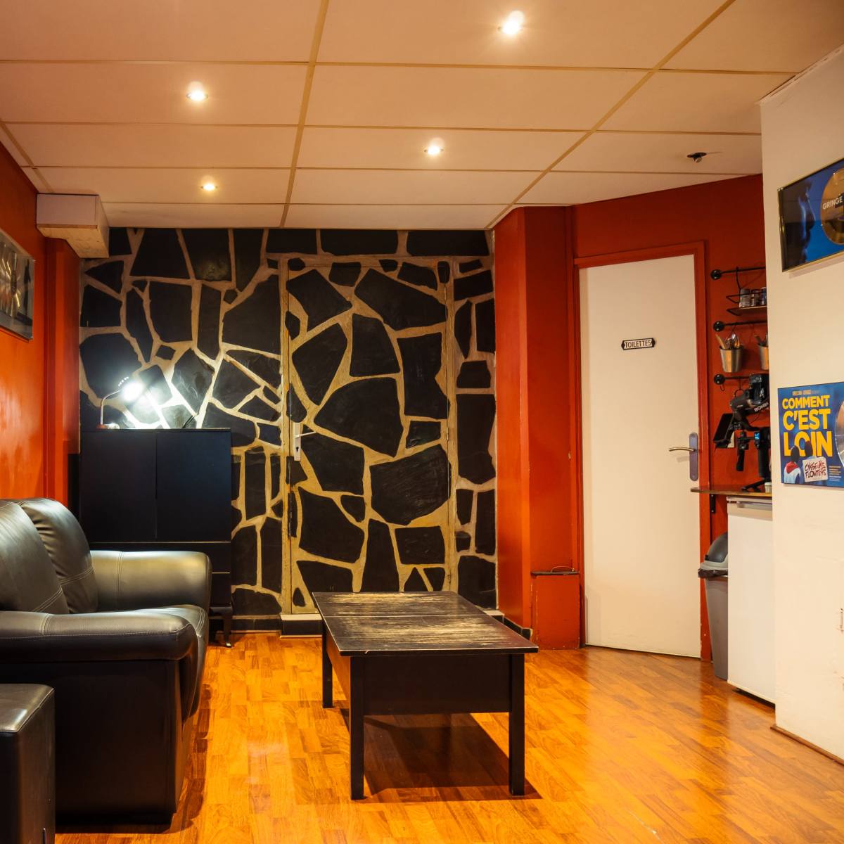 A room with an orange wall, a stone-patterned accent wall, a black leather couch, a wooden coffee table, and a door on the right.