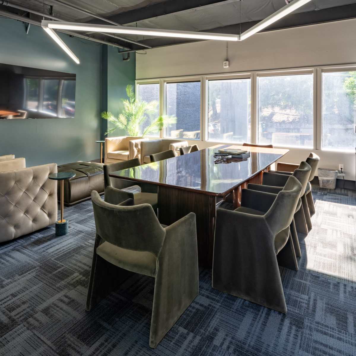 A modern conference room features a large table surrounded by upholstered chairs and large windows providing natural light.
