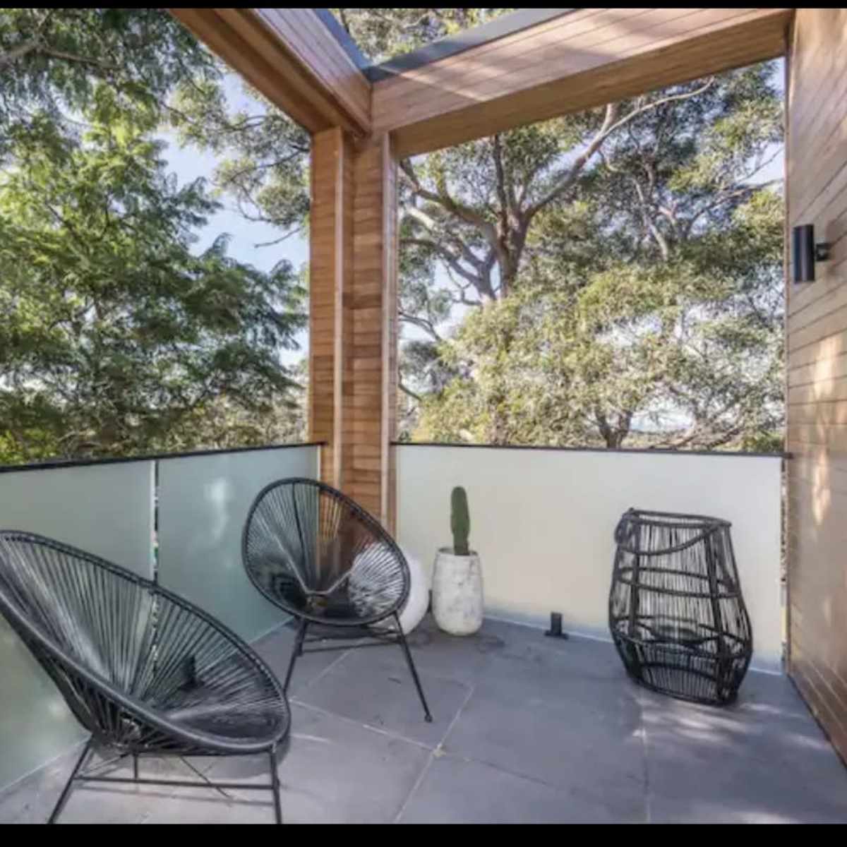A modern balcony with two black chairs, a potted cactus, and a decorative basket, surrounded by greenery.