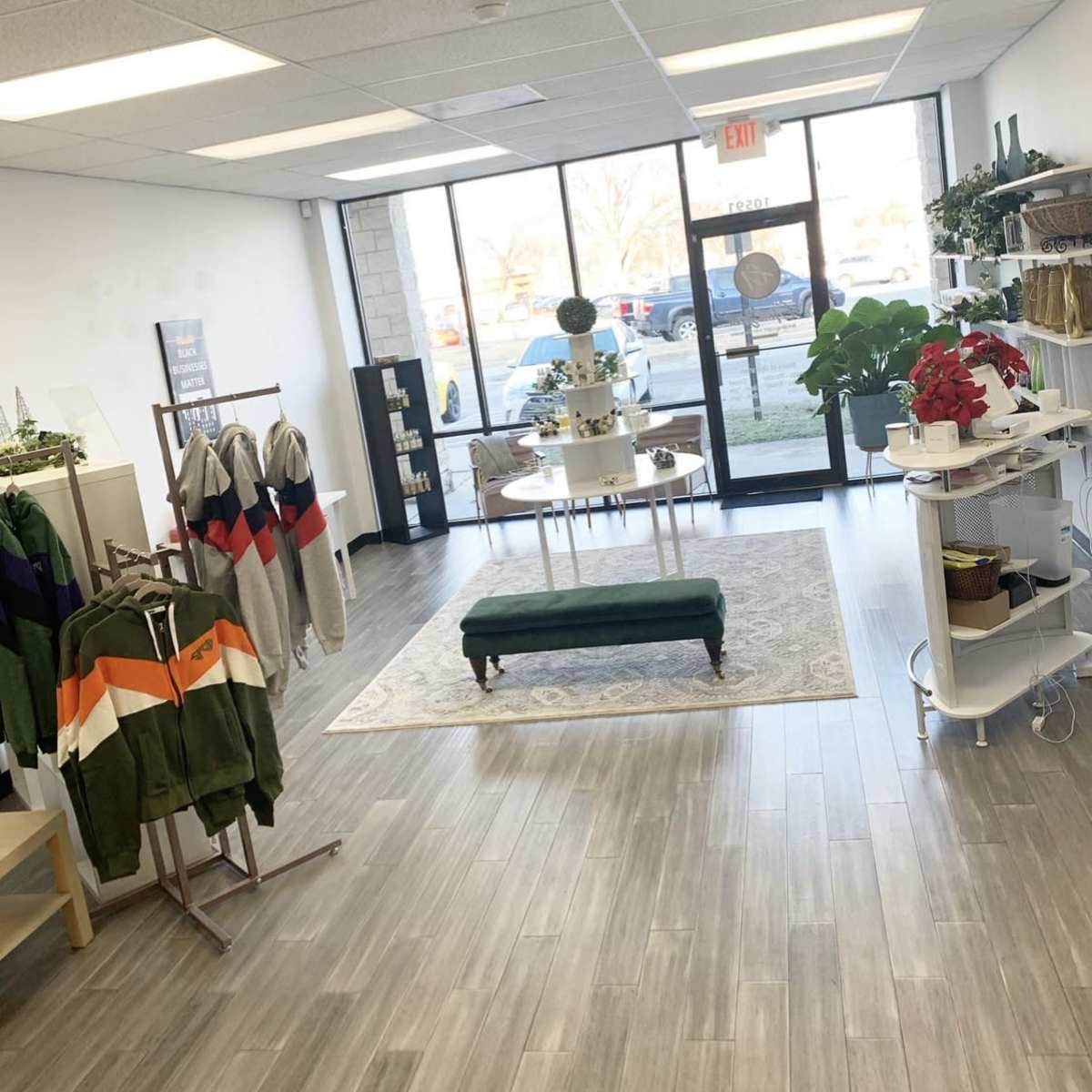 A retail space featuring clothing displays on racks, a decorative table with a cake, and a green bench, with large windows overlooking a parking lot.