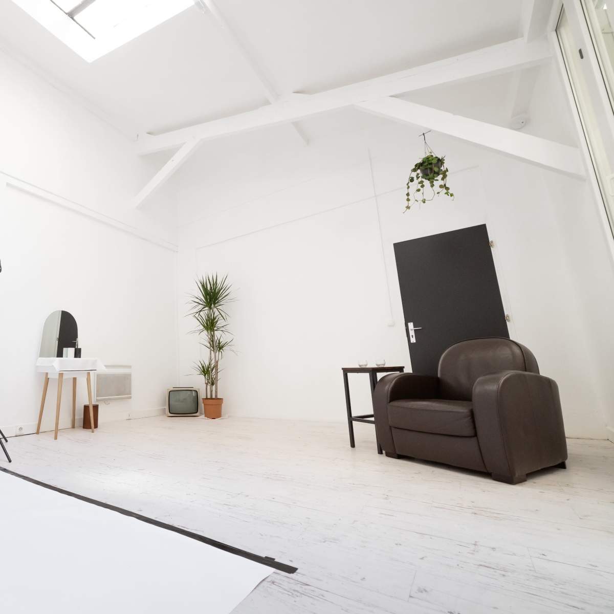 A bright, minimalistic room features a brown armchair, a small table with a mirror, a potted plant, and a softbox light setup against white walls.