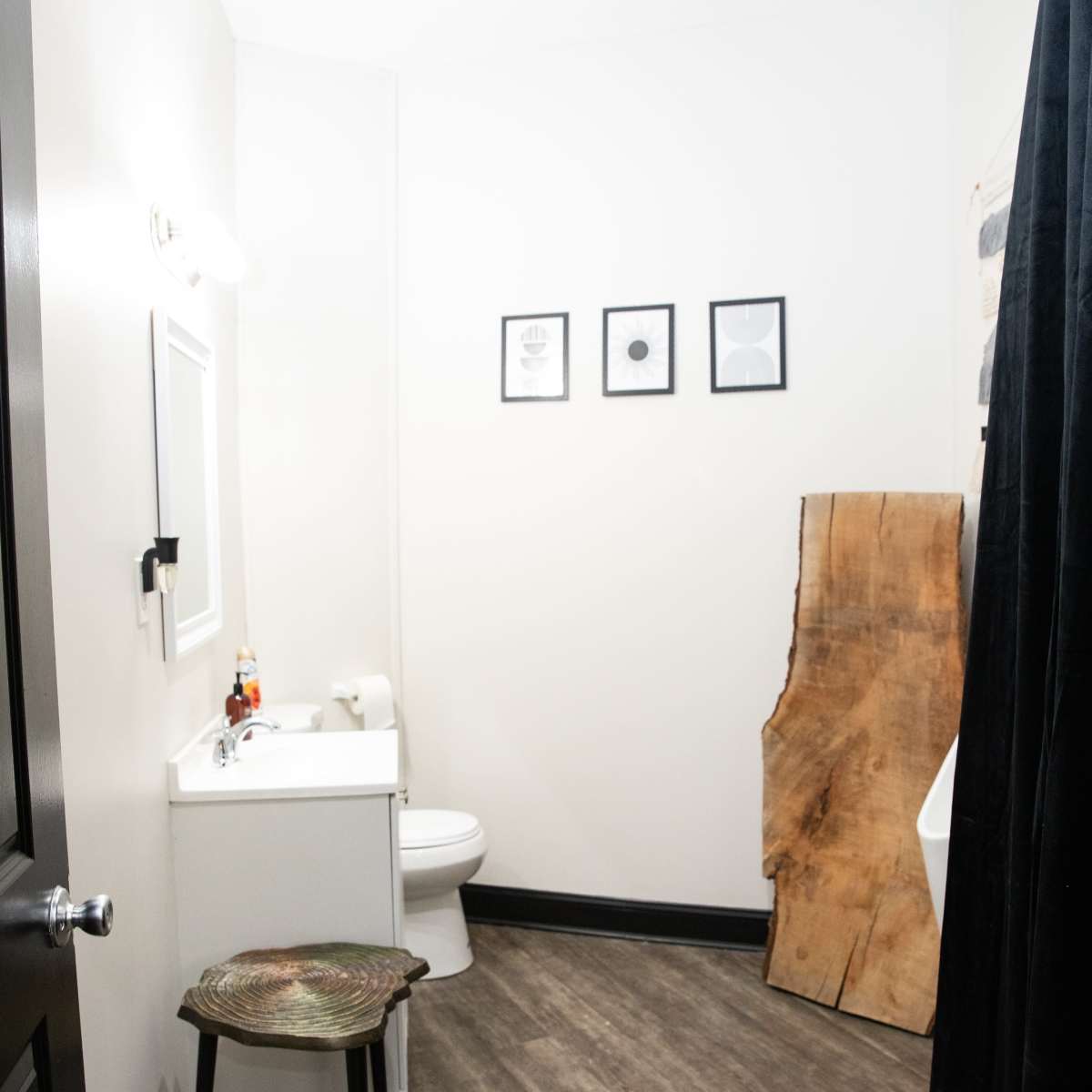 A small bathroom with a toilet, a sink, a framed mirror, four framed pictures on the wall, and a wooden accent piece next to the sink.