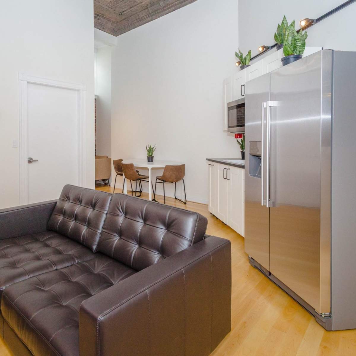 A modern kitchen and seating area feature a black leather couch, stainless steel refrigerator, and a dining set in the background.