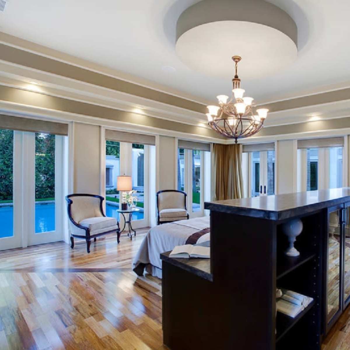 A spacious bedroom with a central chandelier, large windows overlooking a pool, and a wooden floor.