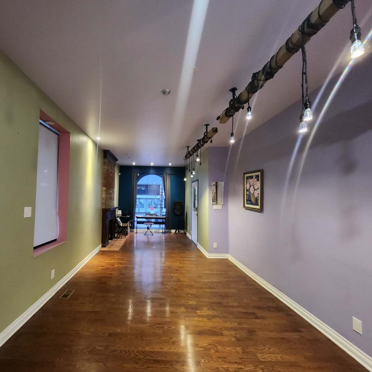 A long hallway with wooden flooring, colorful walls, and track lighting, leading to a window at the far end.