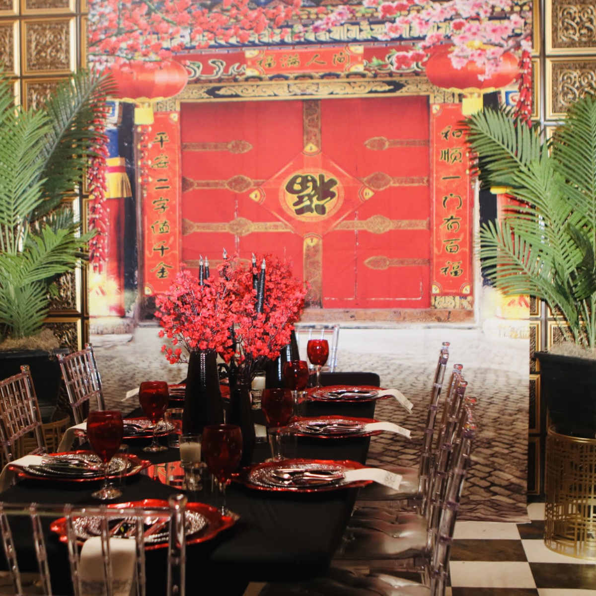 A dining table is set with black and red dinnerware in front of a vibrant mural depicting a traditional red door adorned with flowers and lanterns.