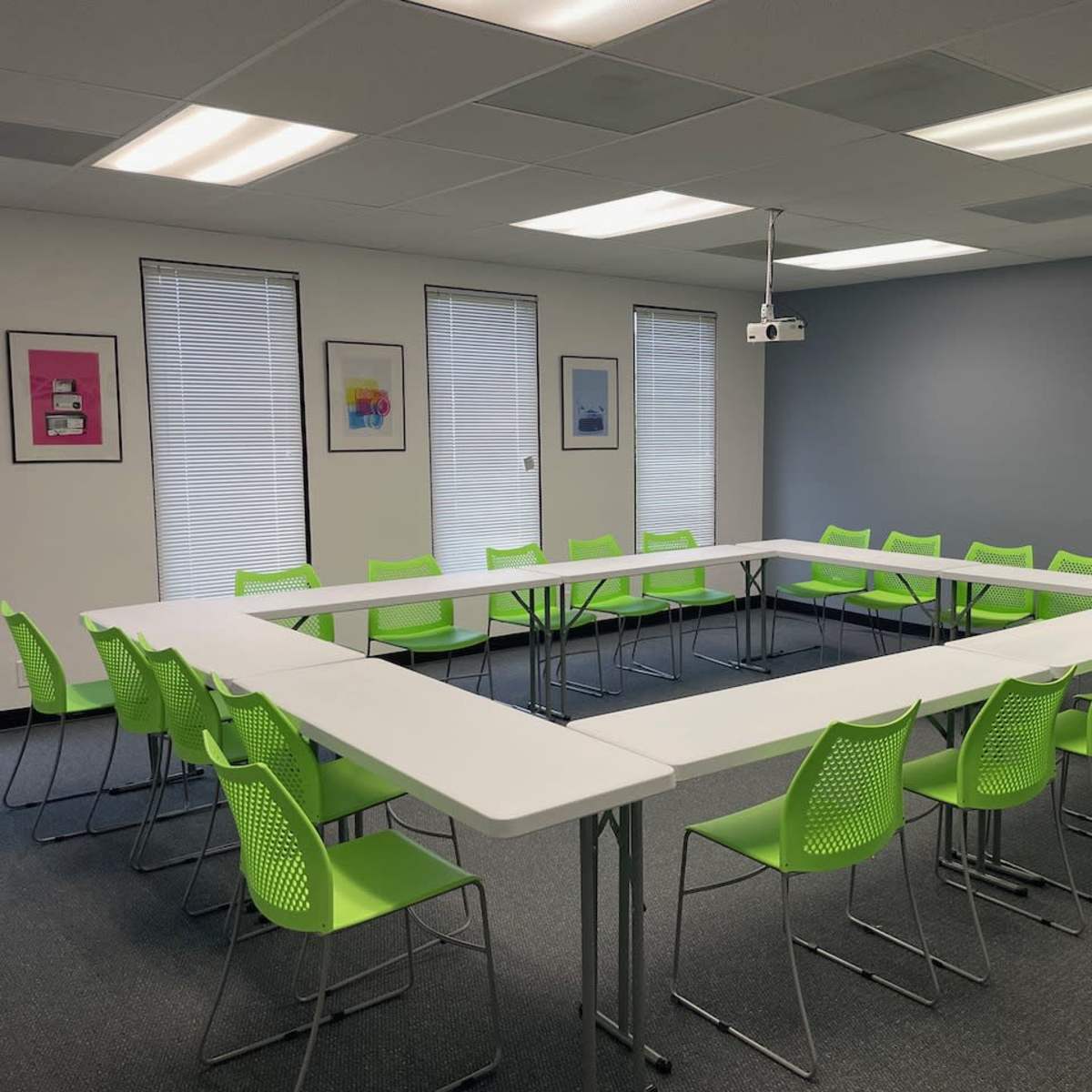 A conference room features a triangular arrangement of white tables surrounded by green chairs, with windows and artwork on the walls.