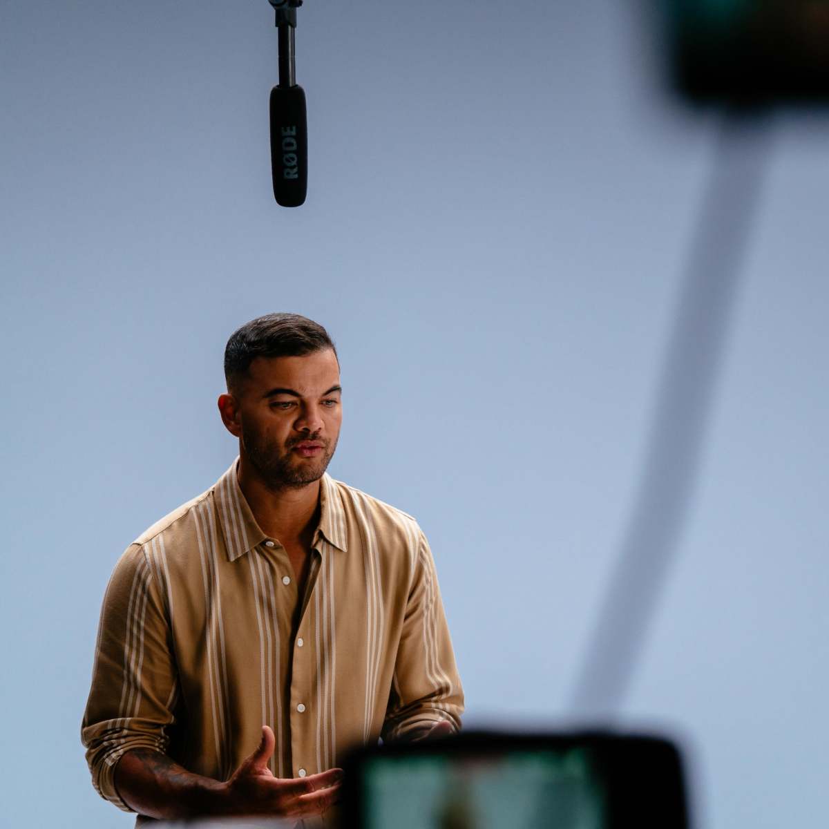 A man wearing a striped shirt speaks while being filmed in a studio.