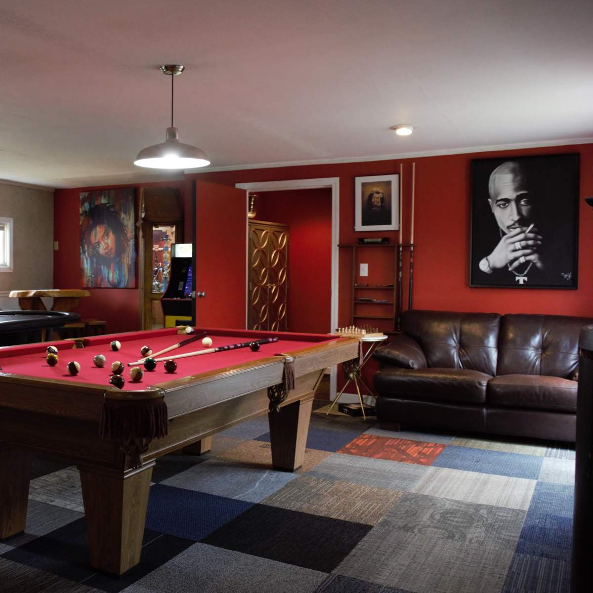 A game room featuring a red-felted pool table, a dark couch, wall art of notable figures, and a flat-screen TV on one wall.