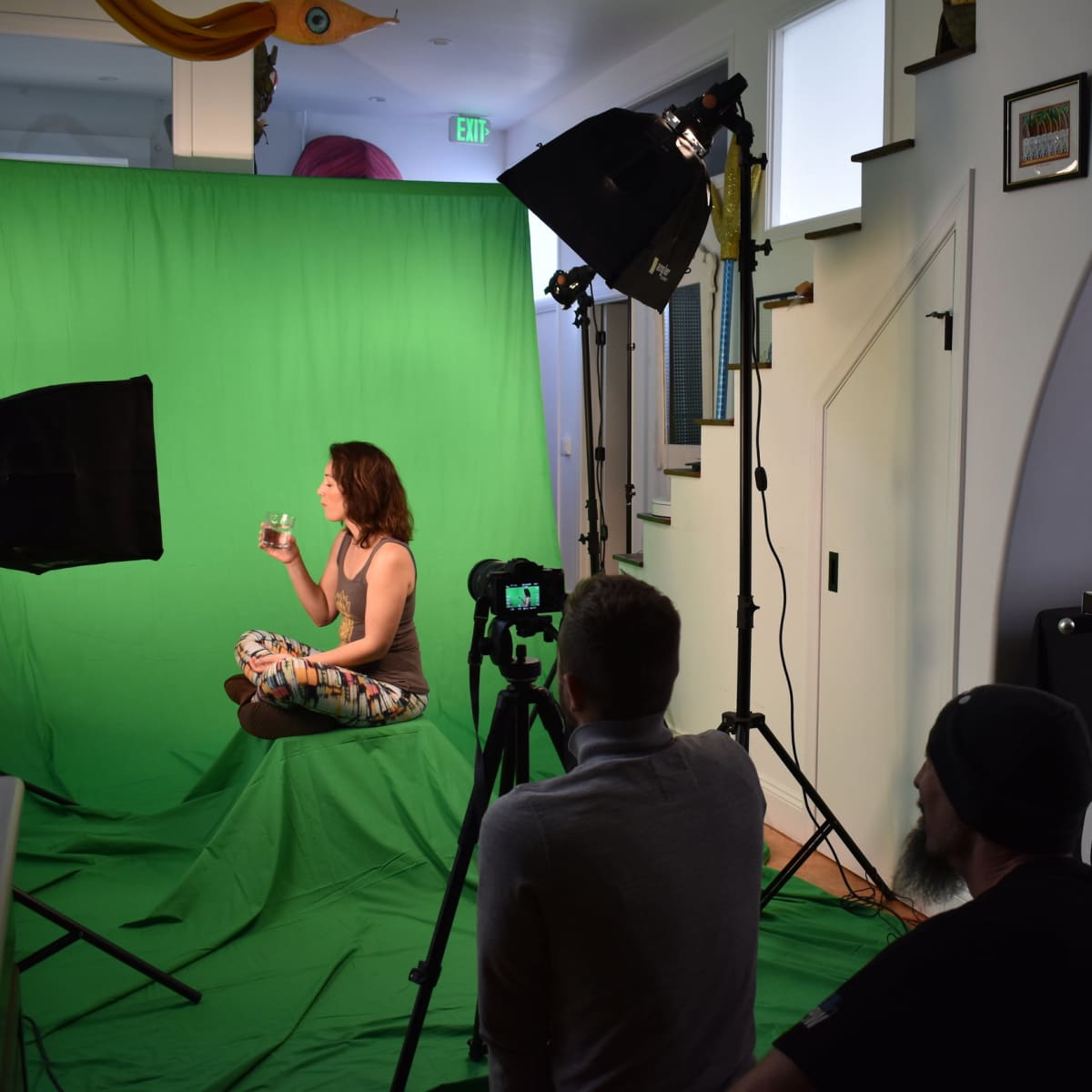 A woman sits cross-legged in front of a green screen while two crew members operate video equipment.