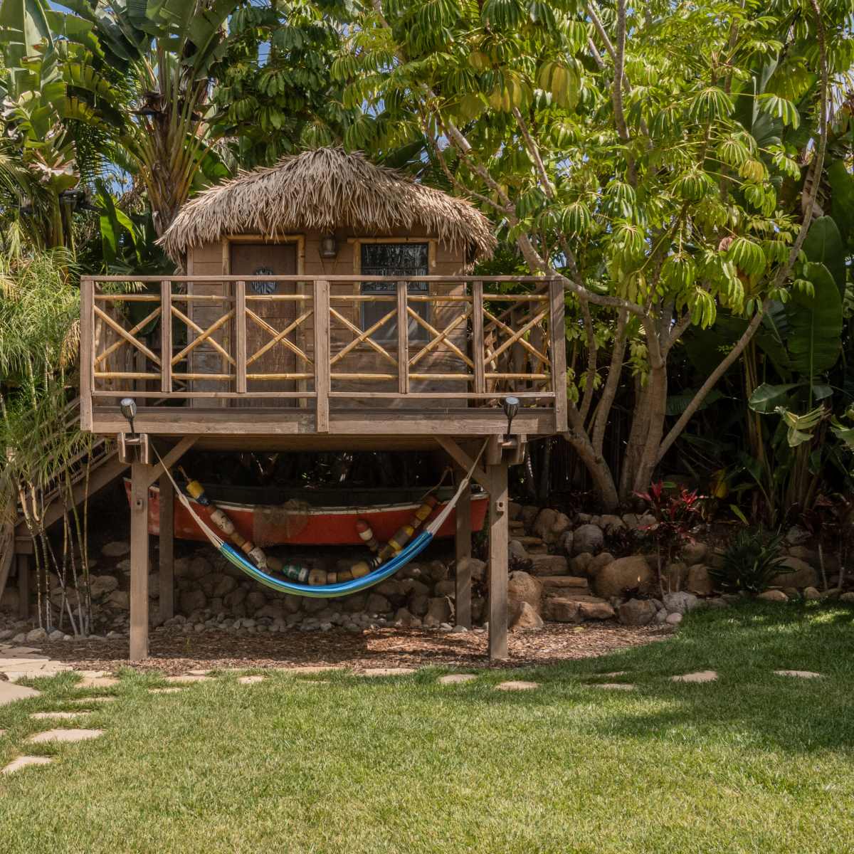 A wooden treehouse with a thatched roof is elevated on stilts, surrounded by tropical plants, and features a hammock suspended below it.