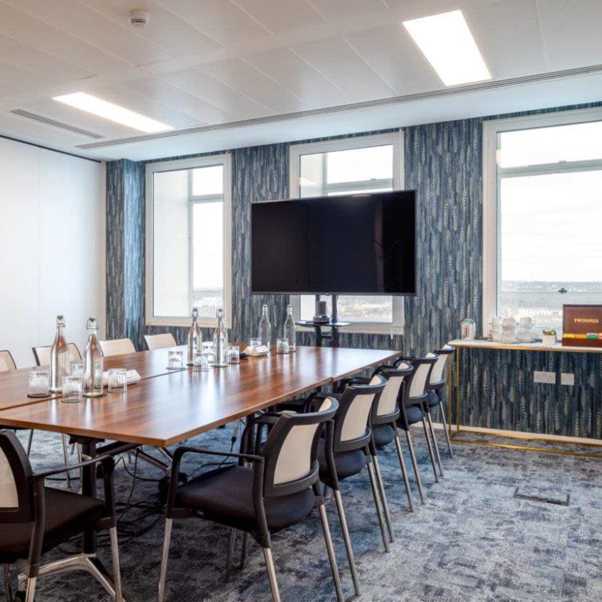 A modern conference room features a large wooden table surrounded by chairs, with a TV mounted on the wall and windows allowing natural light.