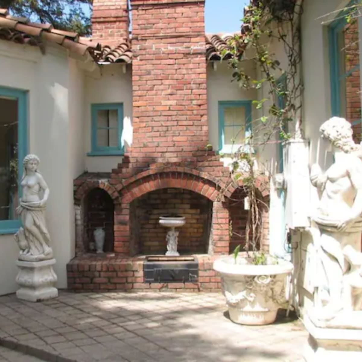 A small courtyard with a brick fireplace flanked by two white statues and surrounded by a white stucco wall and blue window frames.