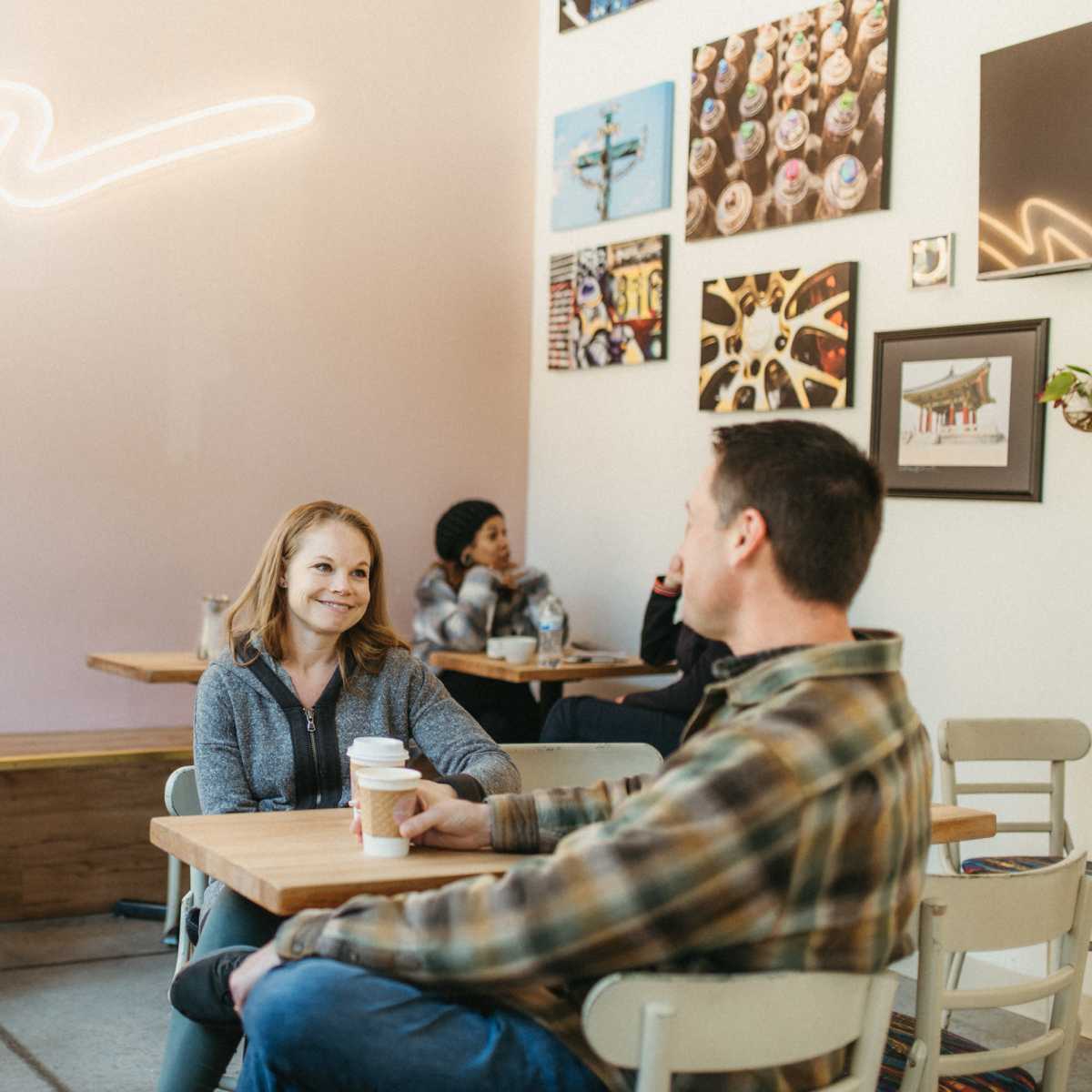 Two people sit at a table in a cafe, engaging in conversation, while other customers are seen in the background.