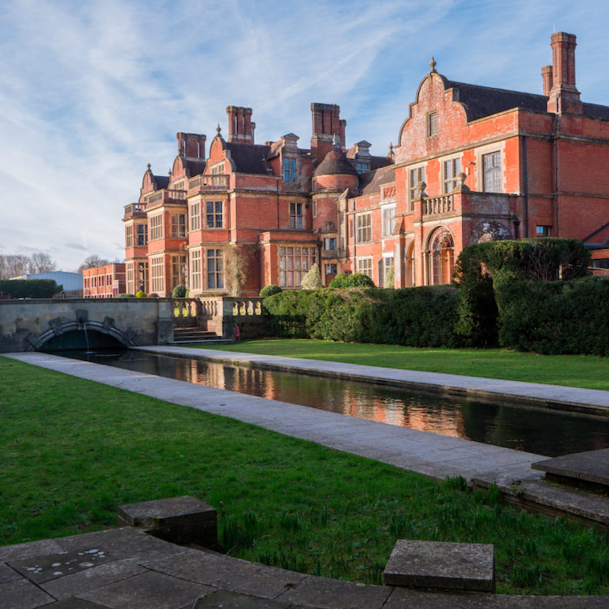 A large red-brick mansion surrounded by neatly trimmed grass and a reflecting water channel in the foreground.