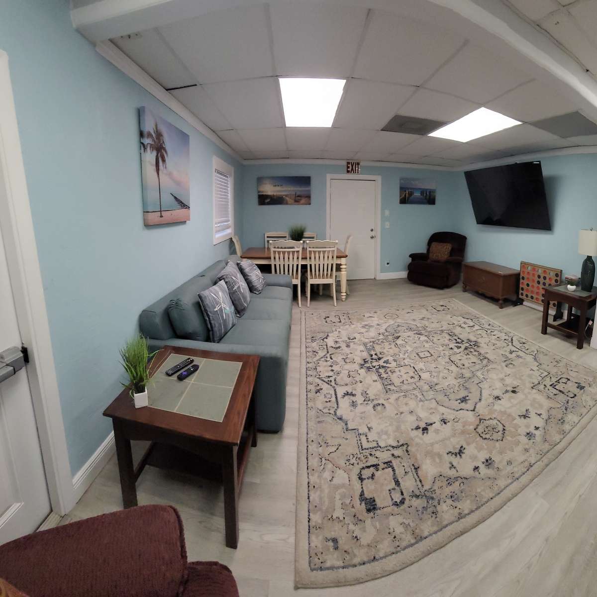 A spacious, light-colored room with a seating area, dining table, and a large television on the wall.