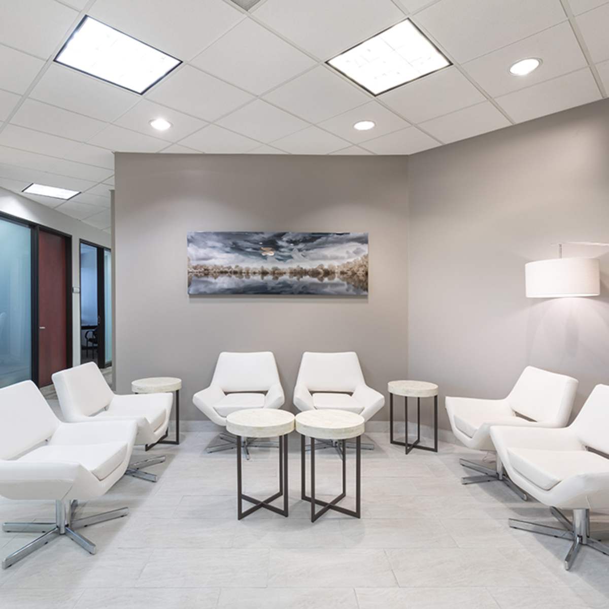 A modern waiting area features white chairs arranged around small tables, with a large painting on the wall and prominent lighting overhead.
