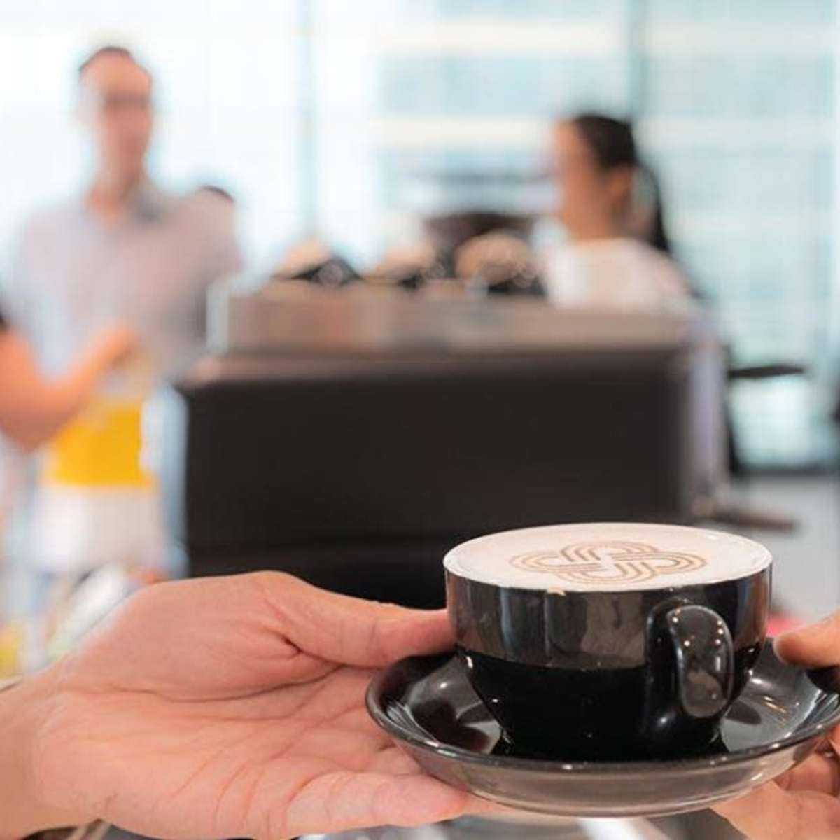 A person hands a black coffee cup with a decorative design on top to another person in a busy café setting.