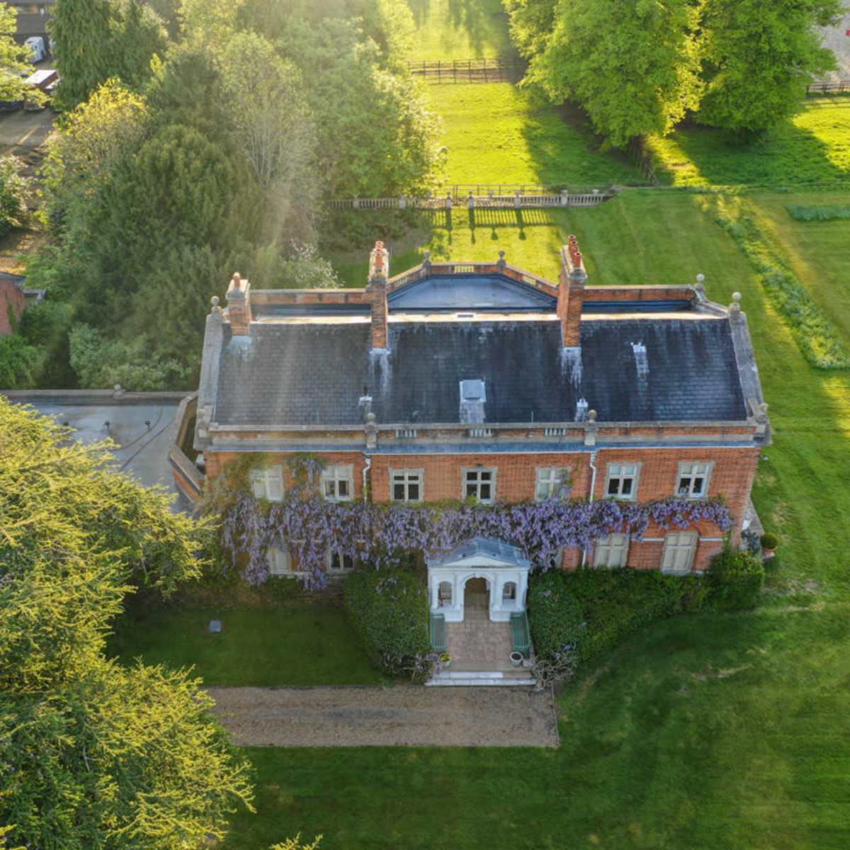 An aerial view shows a large brick house with a white entrance, surrounded by trees and a green lawn.