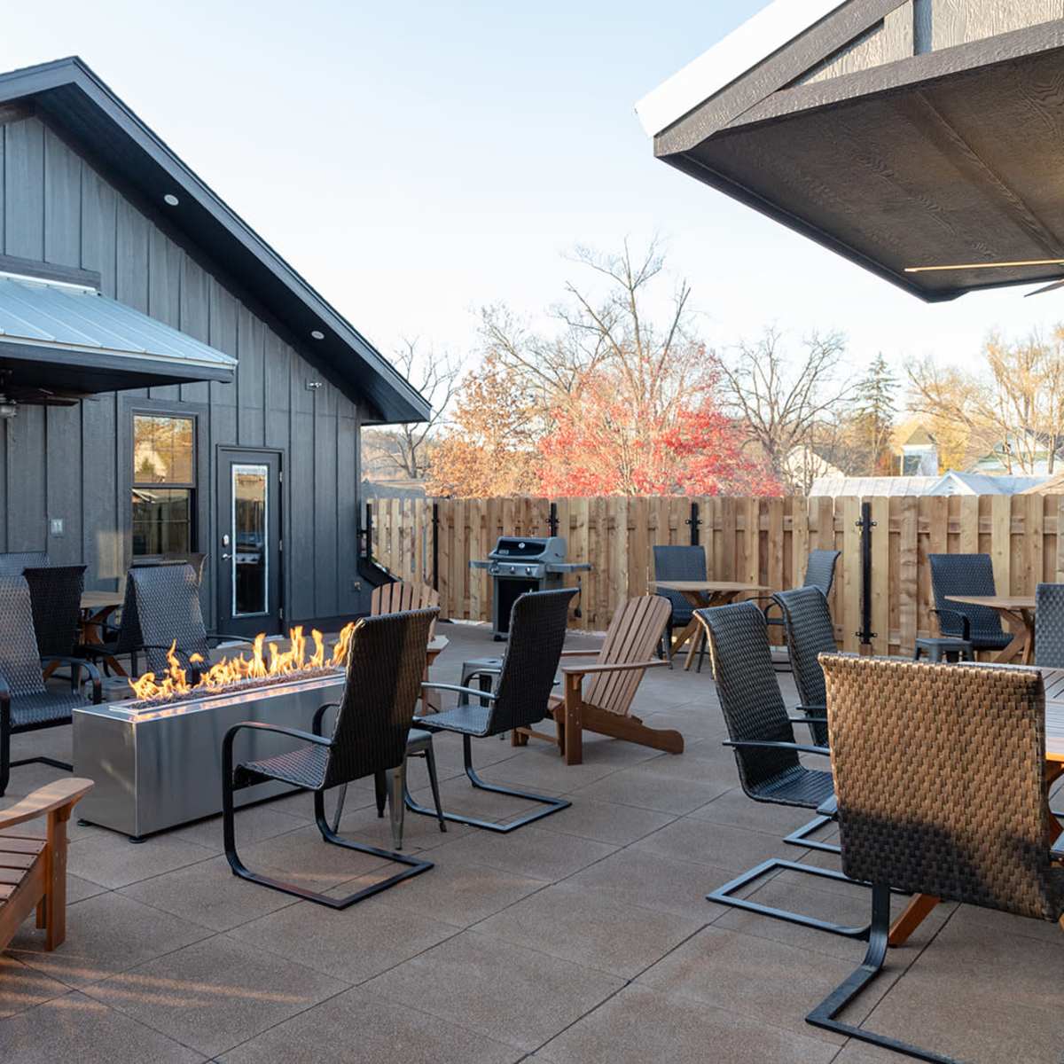 A patio area with several outdoor chairs arranged around a central fire pit, adjacent to a wooden fence and a building in the background.