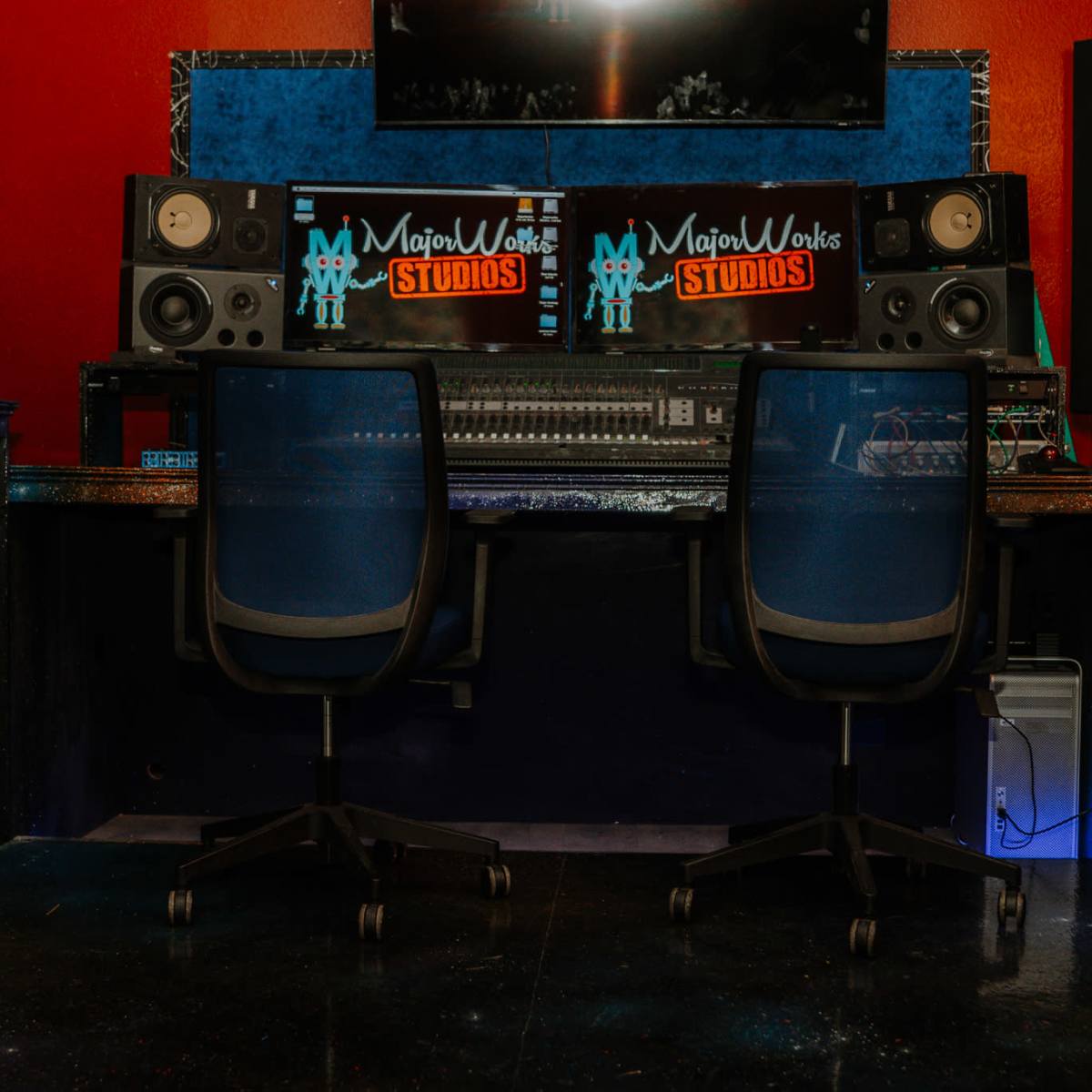 A recording studio with two empty chairs facing a sound mixing console, flanked by large speakers and a screen displaying "MajorWorks Studios."