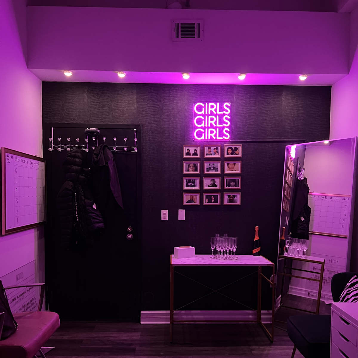 A brightly lit room features a purple neon sign that reads "GIRLS GIRLS GIRLS," a coat rack, a mirror, and a table with champagne glasses.