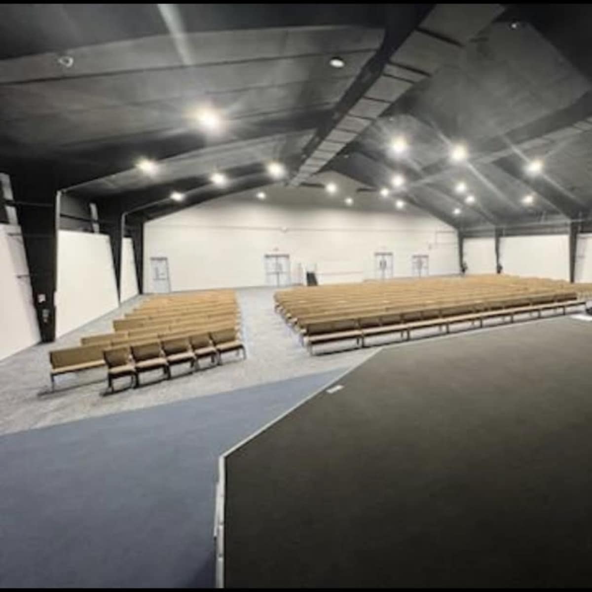 A spacious interior of a hall features rows of wooden benches on a carpeted floor, under bright overhead lighting.