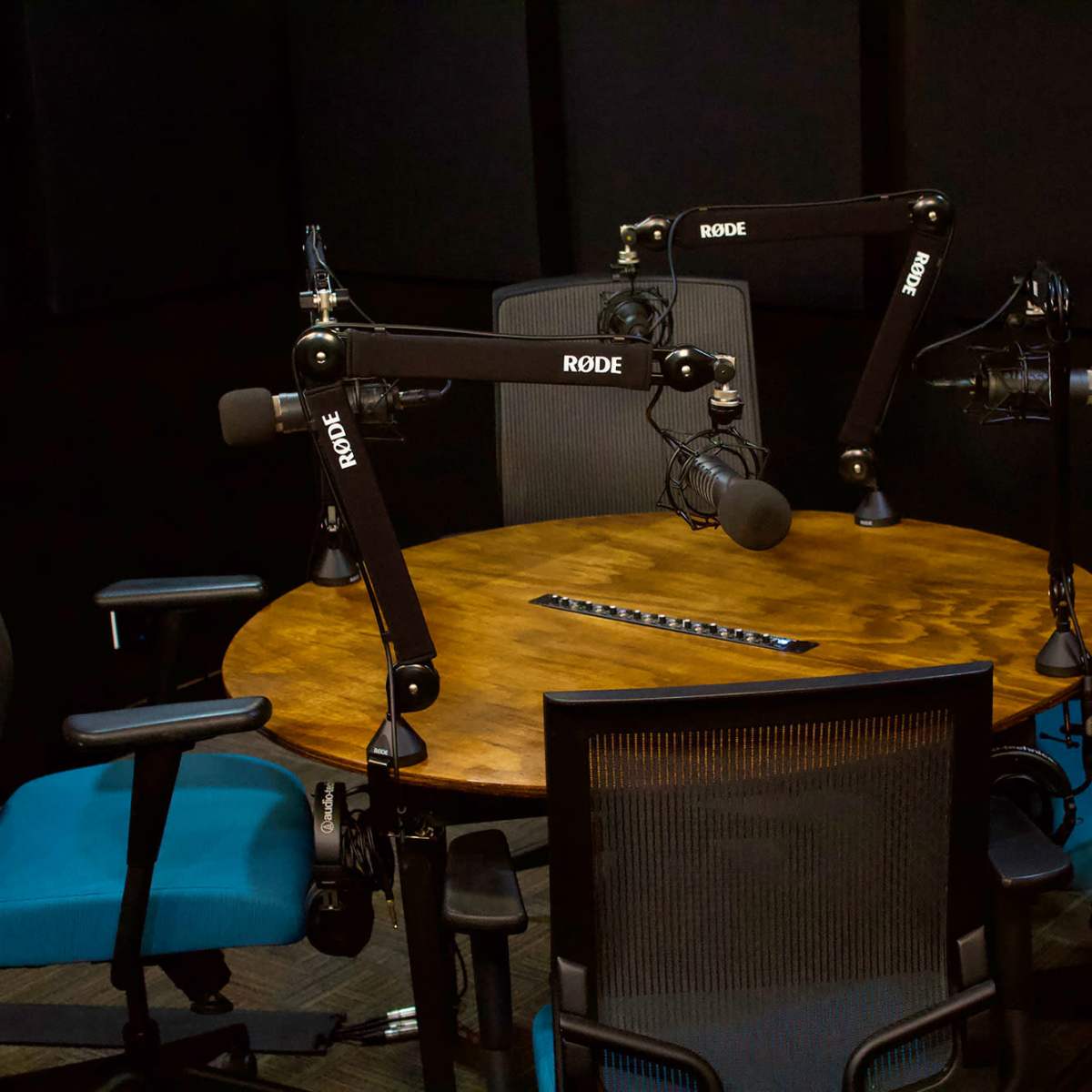 A small podcasting or broadcasting studio featuring a round wooden table with four chairs and adjustable microphone arms mounted overhead.