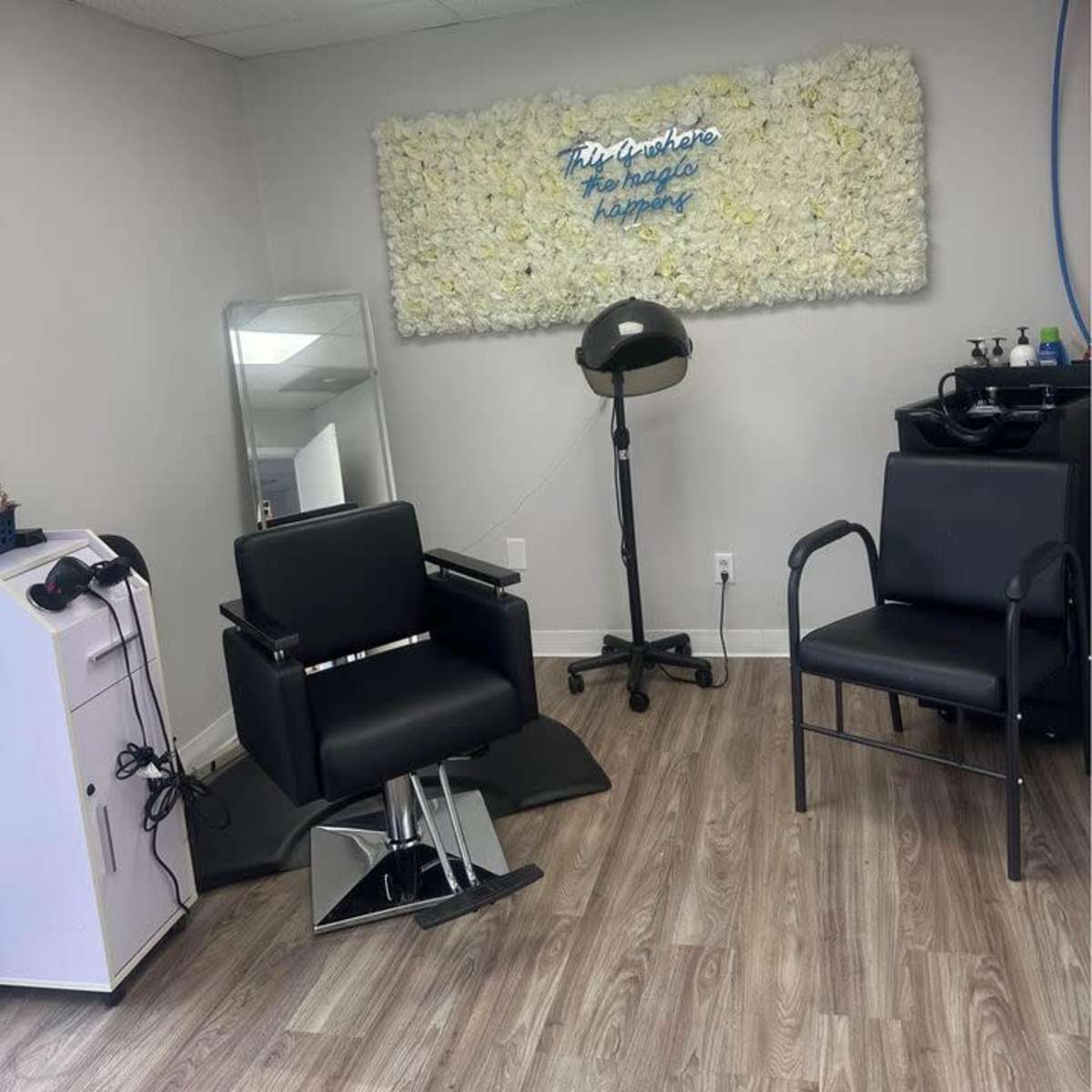 A salon interior featuring two black chairs, a barber mirror, and a wall adorned with a floral arrangement above a sign.
