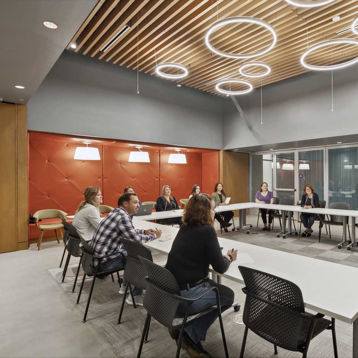 A group of people is seated around a long table in a modern conference room, while one person stands at the front presenting with a podium.