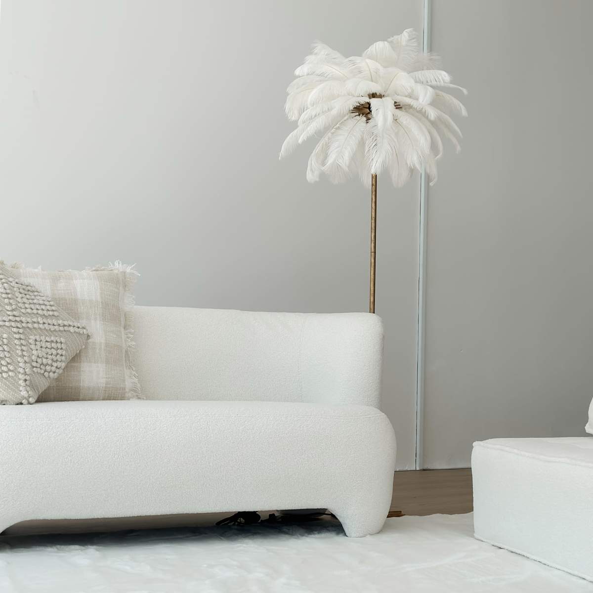A white sofa and a matching ottoman are positioned next to a decorative feather floor lamp against a light gray wall.