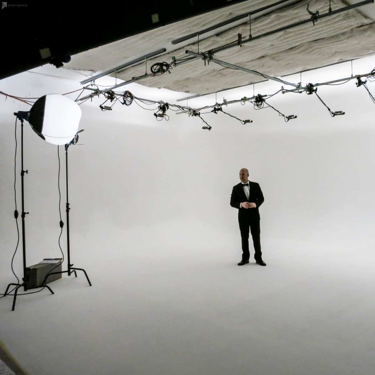 A man in formal attire stands alone in a sparsely populated photography studio with white walls and various lighting equipment.