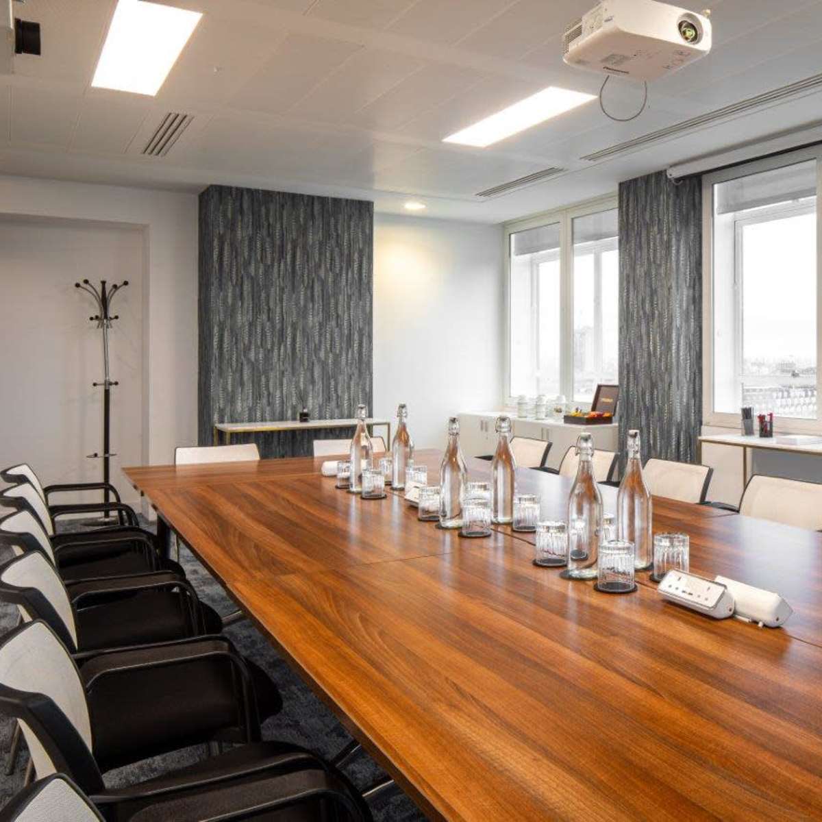 A modern conference room features a long wooden table surrounded by chairs, with a projector and large windows overlooking a cityscape.
